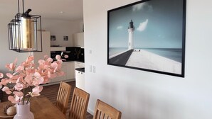 Ein Essbereich mit Holztisch, Stühlen, einer Vase mit rosa Blumen, einer Pendelleuchte und einem gerahmten Bild eines Leuchtturms an der Wand.
