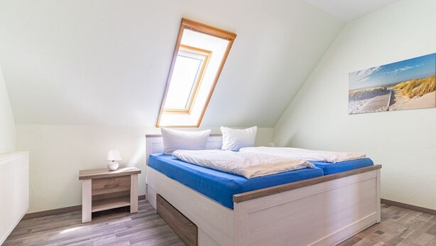 Een slaapkamer met een schuine plafond, dakraam, bed met blauw beddengoed, nachtkastje met een lamp, en een schilderij aan de muur.