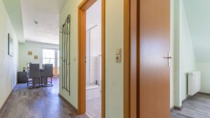 Een gang met een houten deur, een eetgedeelte met stoelen, en een zicht op een badkamer.