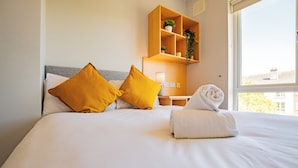 Una habitació d'hotel amb un llit, dos almoinos, una taula de nit amb una làmpada, una prestatgeria amb plantes i una finestra.