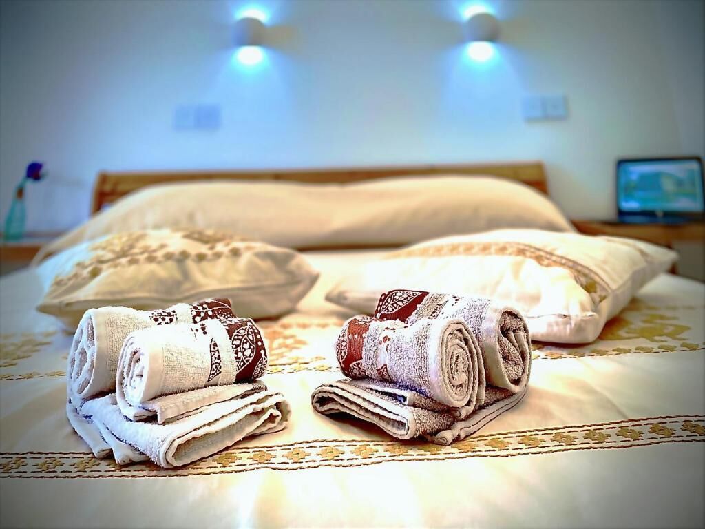 Una cama bien hecha con toallas enrolladas y un almohadón, una mesita de noche con lámpara, y un televisor al fondo.