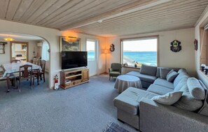 A living room with a television, a sofa, a dining table, and a view of the sea.