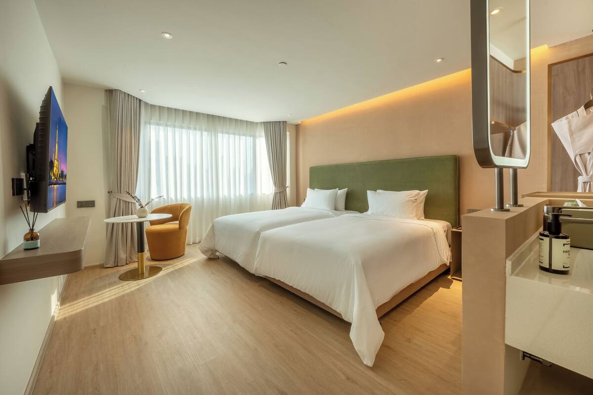 A modern hotel room with a large bed, a sitting area with a chair, a small table, a TV, and a mirror.