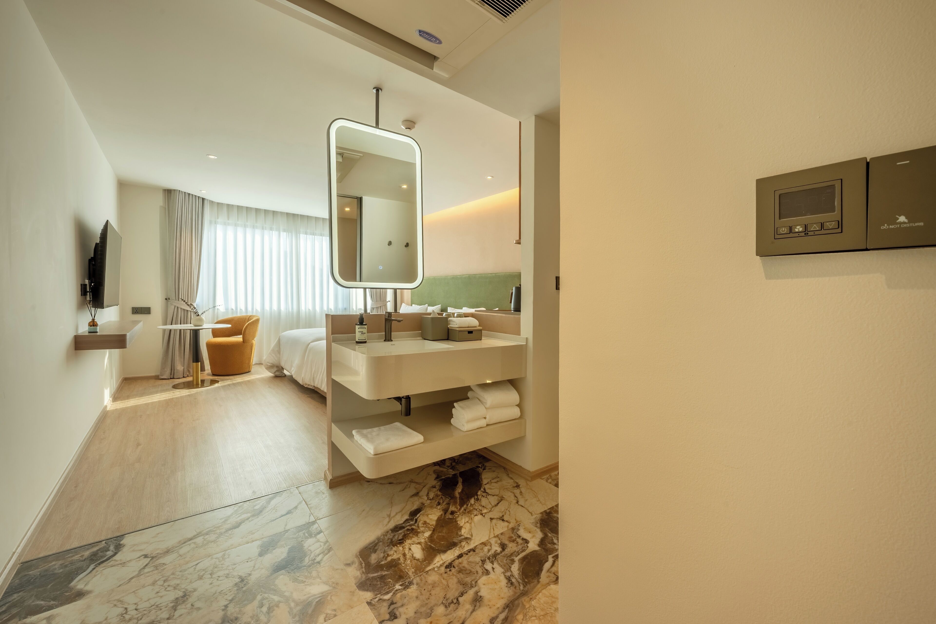 A modern hotel room with a large mirror, a built-in TV, a vanity with a sink, and a seating area with a chair and table.