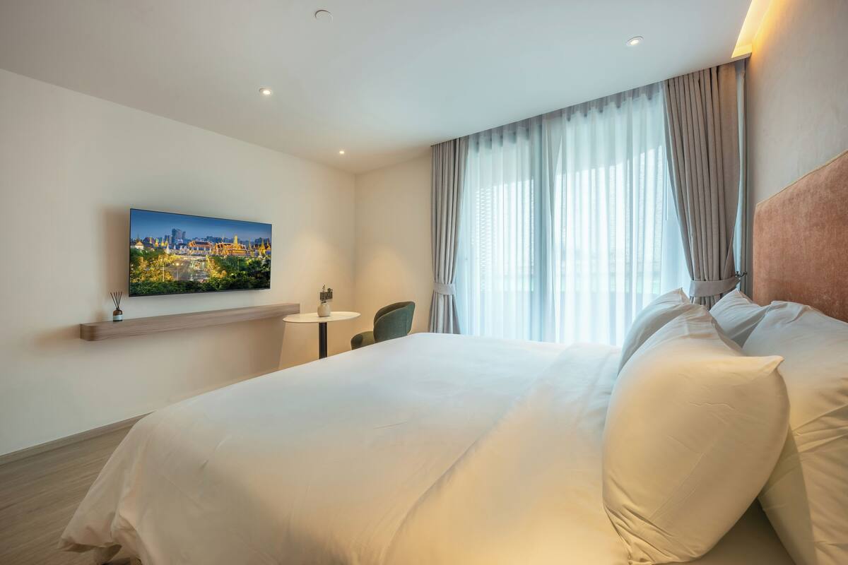 A hotel room with a large bed, a small table, a chair, a painting on the wall, and a window with curtains.