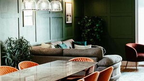 Un espace de salle à manger moderne avec une longue table et des chaises orange, un mur vert et une grande plante.