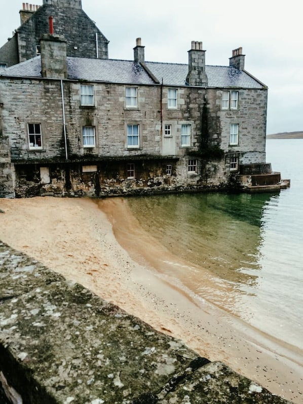 A stone building with multiple chimneys and windows, adjacent to a calm body of water. - NEW to VRBO: Luxury Seafront Stay in Central Lerwick W/ Parking (Lerwick)