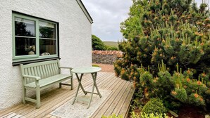A wooden deck with a bench and table, a white building with a window, and a coniferous tree with orange cones.