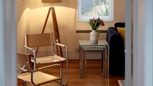 Una habitación con piso de madera, una lámpara de pie, una silla, una mesita con un florero y una ventana con persianas.