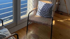 Una habitación con un gran ventanal corredero que da a una vista de agua y un pueblo. Hay una silla con un cojín estampado y una lámpara de pie sobre el suelo.