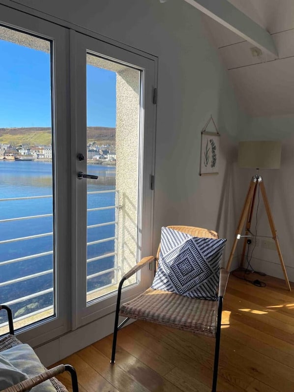 A room with a large sliding glass door leading to a view of a body of water and a town. There is a chair with a patterned cushion and a tripod lamp on the floor.