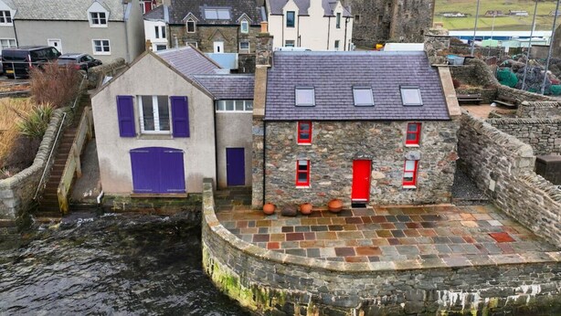 Un edificio de piedra con puerta roja, una ventana pequeña y un muro de piedra.