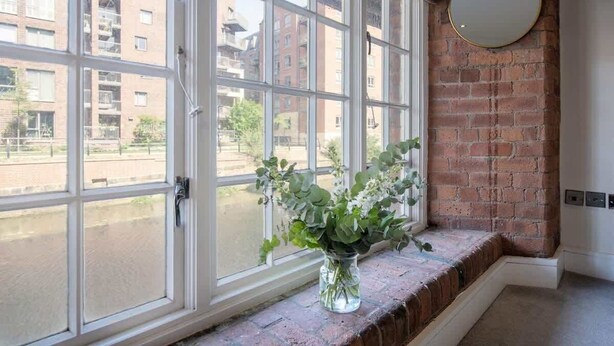 A room with a large window, a vase with flowers, a brick wall, and a round mirror. - The Cacao Suite - Charming River View Apartment Steps from York City Centre (York)