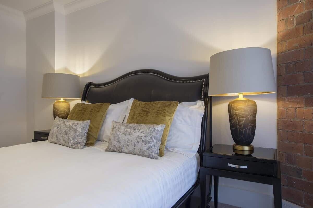 A neatly made bed with white linens and gold and white pillows, flanked by two bedside tables with lamps.