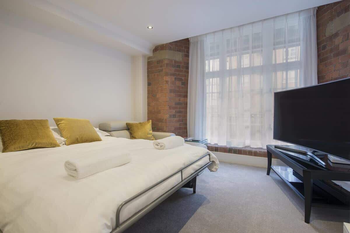 A hotel room with a large bed, a flat-screen TV, a brick wall, and a window with sheer curtains.