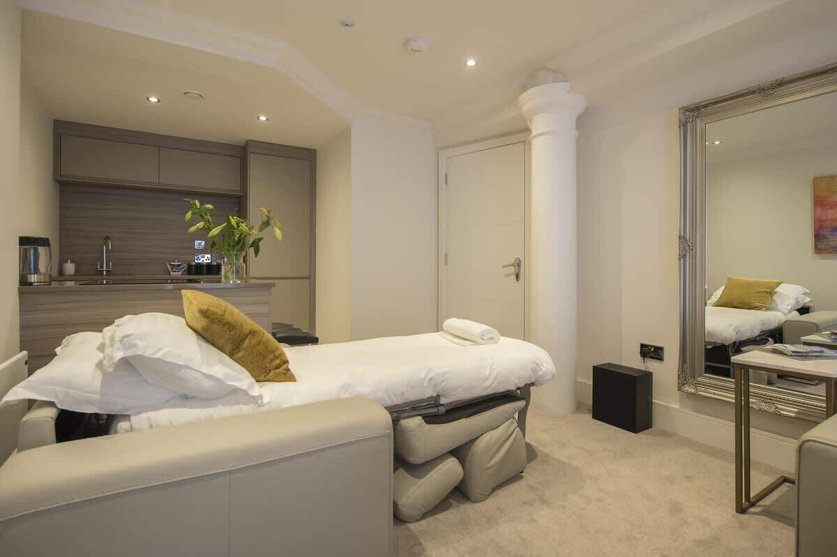 A modern bedroom with a bed, a bedside table, a mirror, and a small kitchenette area.