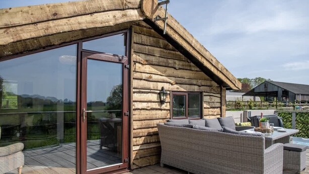 Een vakantiehuis van hout met een glazen deur die uitkomt op een terras, rieten meubels en uitzicht op het omliggende landschap.