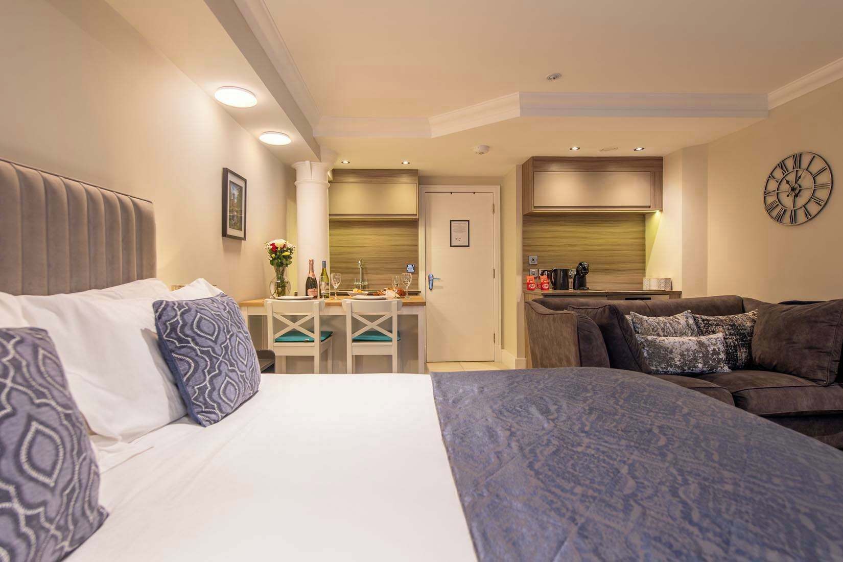 A hotel room with a bed, a sofa, a dining table with chairs, a kitchen area, and a clock on the wall.