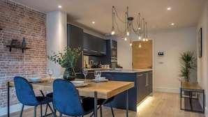 A modern kitchen with a brick wall, dark cabinets, and a wooden dining table with blue chairs. - The Botanical Suite - Luxury Apartment with Balcony & York Minster View (York)