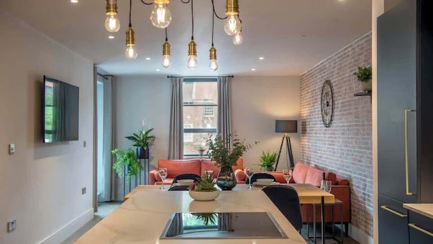 A modern kitchen with a central island, a dining area with a sofa, and a brick wall with a clock. - The Botanical Suite - Luxury Apartment with Balcony & York Minster View (York)