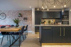 A modern kitchen with dark cabinetry, a central island, and pendant lighting. - The Botanical Suite - Luxury Apartment with Balcony & York Minster View (York)