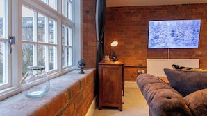 A cozy living room with a brick wall, a large window, a flat-screen TV, a wooden side table, and a cushioned sofa. - The Refinery Suite - Stylish York Apartment in Historic Building (York)