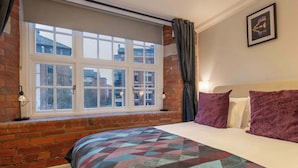 A bedroom with a large window, a brick wall, a bed with patterned bedding, and a framed picture on the wall. - The Refinery Suite - Stylish York Apartment in Historic Building (York)