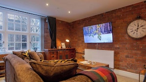 A cozy living room with a brick wall, a large window, a sofa, a TV, and a clock. - The Refinery Suite - Stylish York Apartment in Historic Building (York)