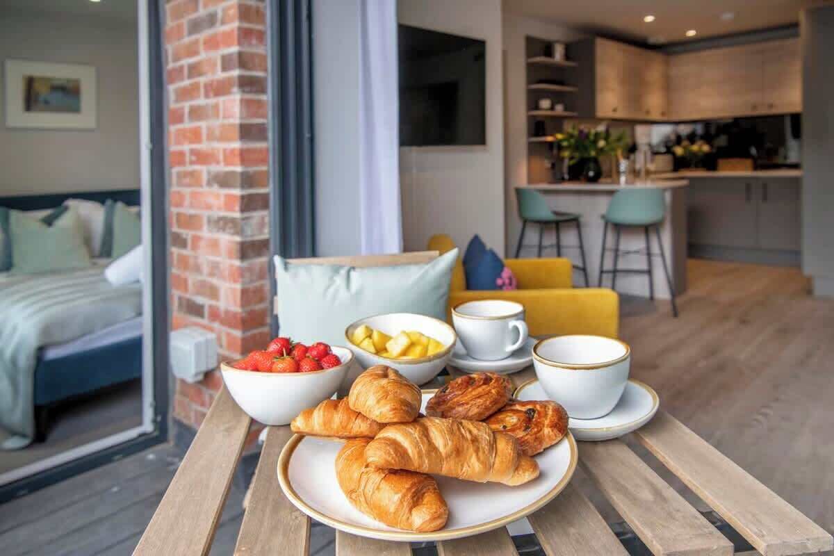 A breakfast setting with croissants, fruit, and coffee on a table in a modern room with a bed, a brick wall, and a kitchen in the background.