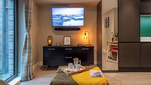 Un salon moderne doté d’un téléviseur à écran plat, d’un meuble de rangement pour appareils électroniques de couleur sombre, d’un coin salon confortable agrémenté d’un plaid jaune et offrant une vue par une fenêtre.