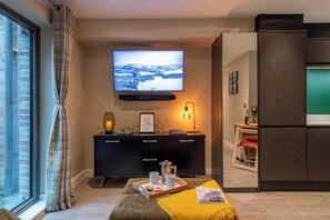 Ein modernes Wohnzimmer mit einem Flachbildfernseher, einer dunkelbraunen Unterhaltungsecke, einer gemütlichen Sitzecke mit einer gelben Decke und Blick durch ein Fenster.