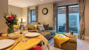 Un coin repas avec une table dressée pour deux personnes, un canapé, une horloge au mur et une porte-fenêtre coulissante donnant sur un espace extérieur.