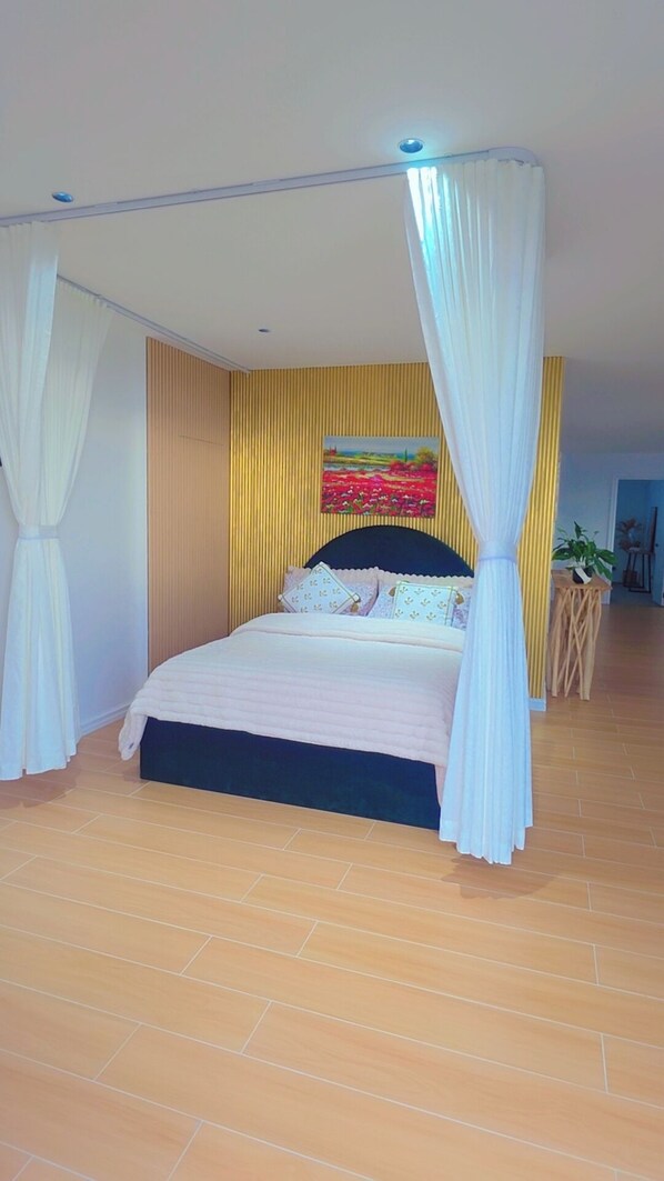 A bedroom with a bed, curtains, a painting on the wall, and a small table with a plant.
