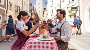 Straßenszene mit Personen, die an Tischen im Freien essen, einem Glockenturm im Hintergrund und Gebäuden, die die Straße säumen.