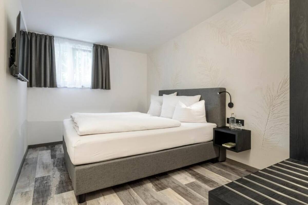 Una habitación de hotel moderna con cama, mesita de noche, televisión y ventana con cortinas.