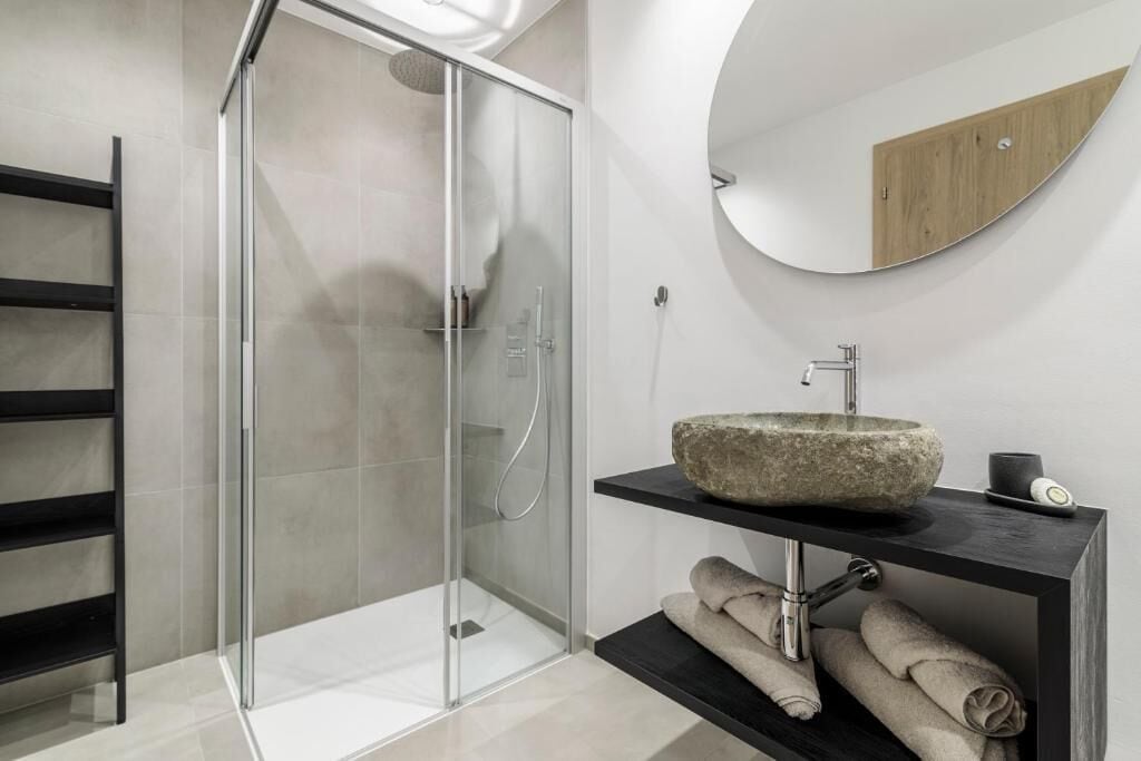 A modern bathroom with a glass shower enclosure, a stone sink, and a wooden shelf with towels.