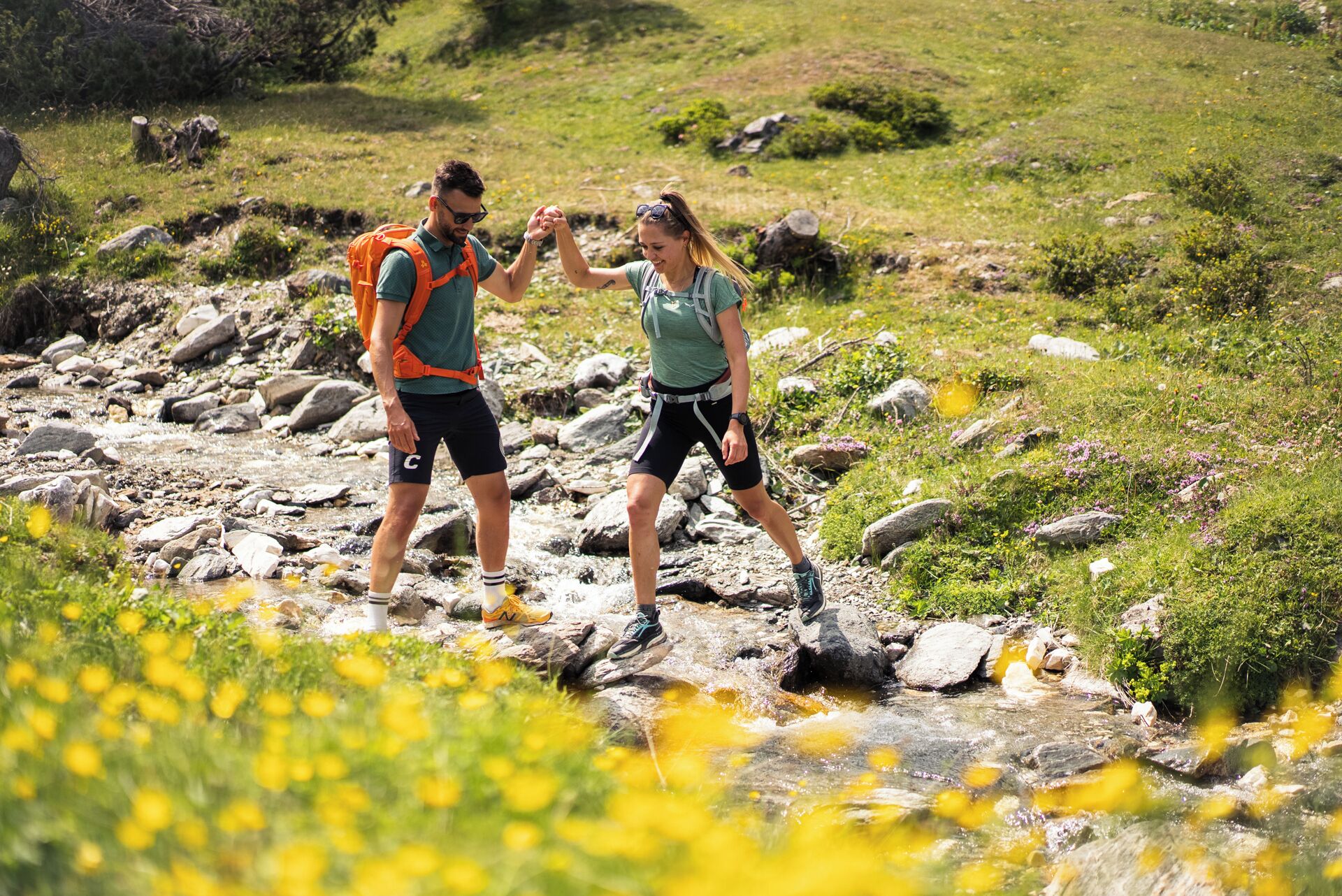 Two people hiking in a rocky, grassy area with a stream.