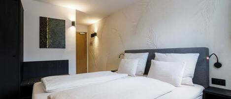 Cama bem arrumada com roupa de cama branca, cabeceira escura e luminária fixada na parede.