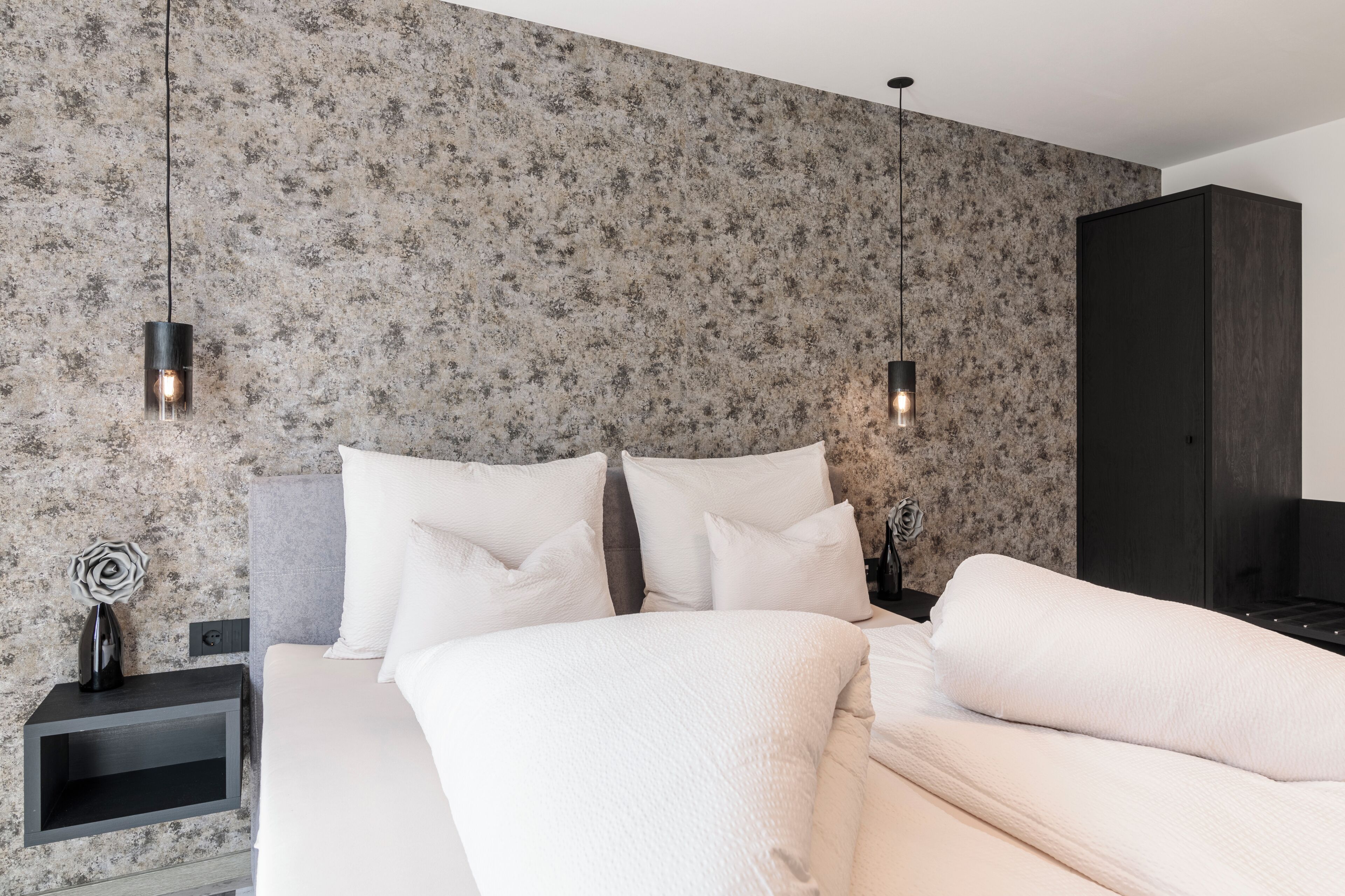 A bedroom with a bed, white pillows, a nightstand, a dresser, and a wall with a textured pattern.