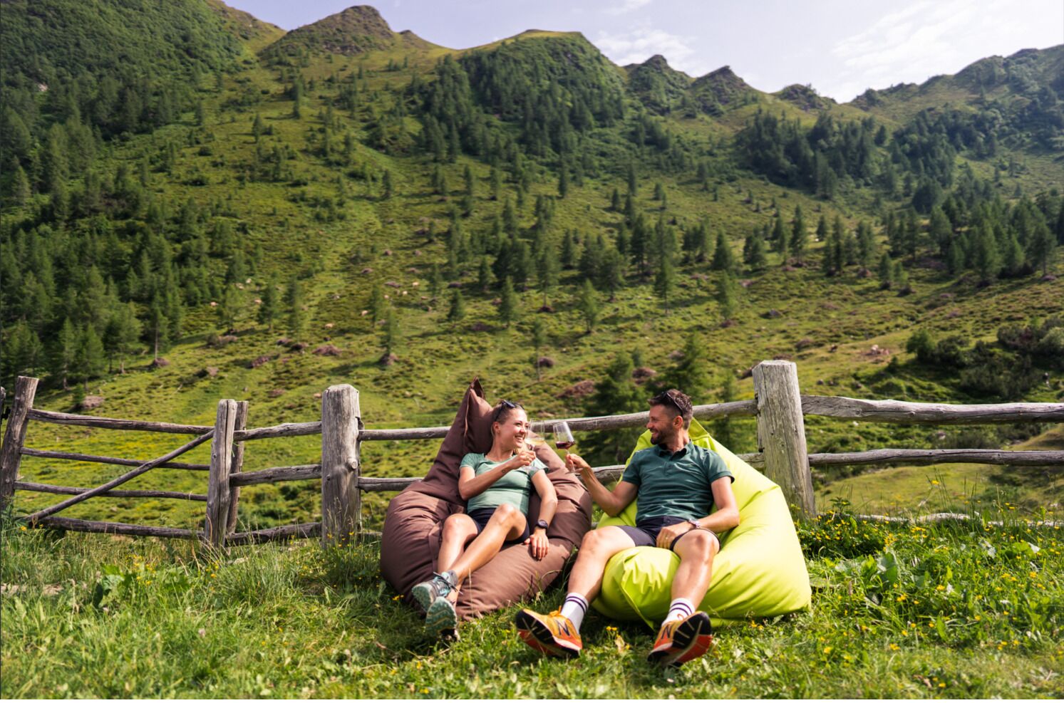 Three people relaxing on bean bags in a grassy field with a wooden fence and mountains in the background.