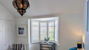 A bedroom with a bed, a window with blinds, a chair, a desk with a laptop, and a decorative lamp. - Central Cute Garden Cottage Close to Town & Beach (Brighton and Hove)