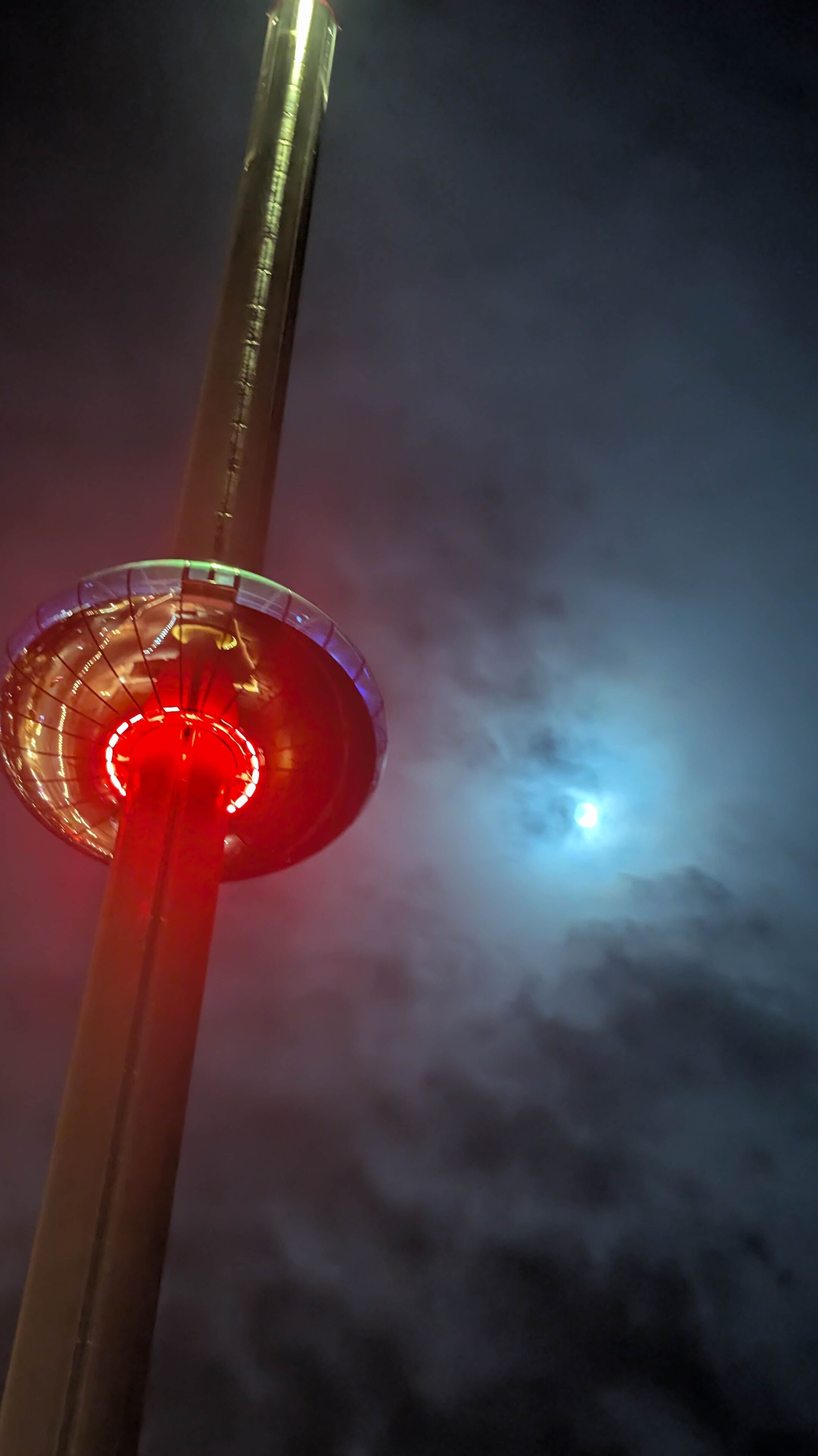 Uma estrutura alta com uma luz circular vermelha no topo, com um céu nublado ao fundo.