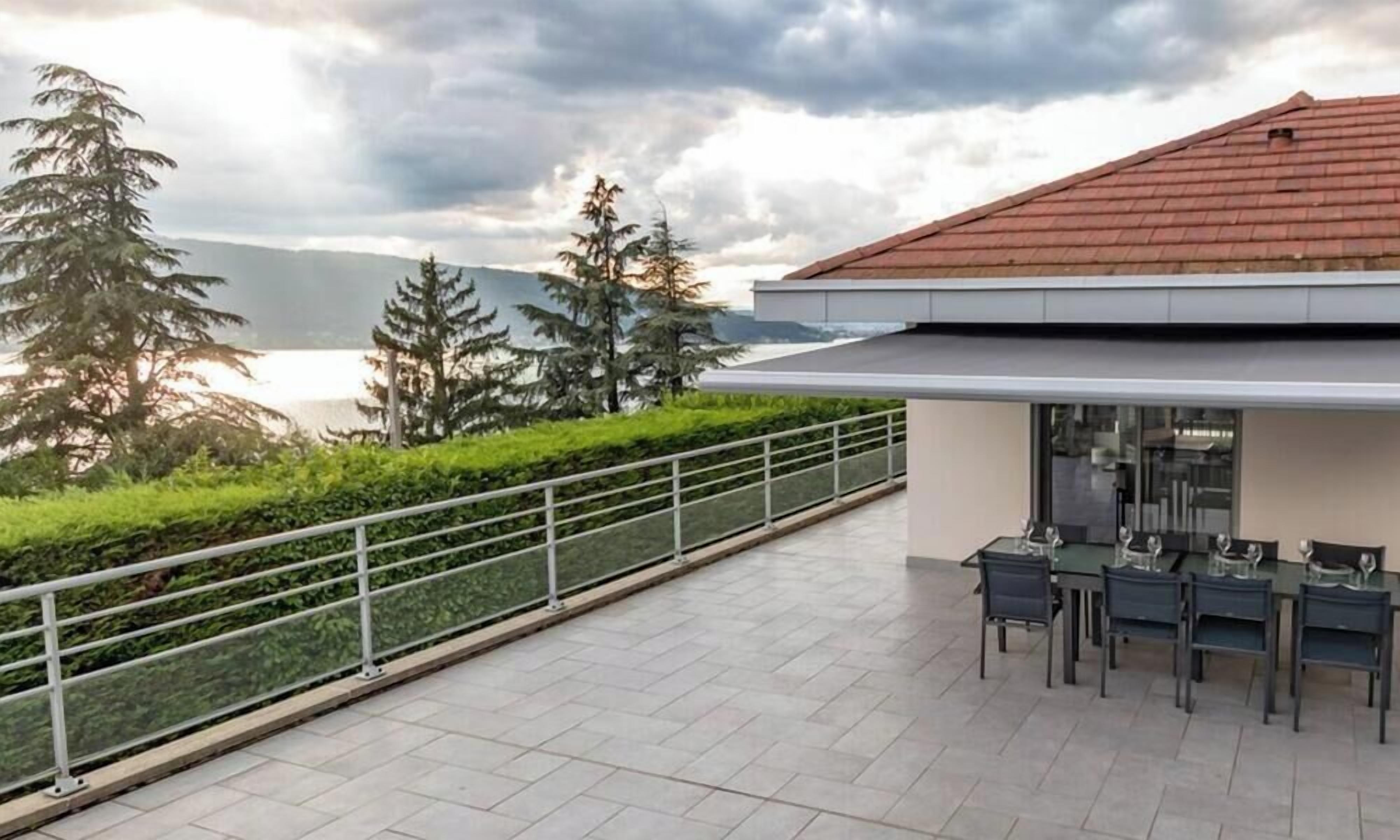 Un bâtiment moderne avec un toit rouge, une terrasse dotée d’une balustrade et un espace repas avec des tables et des chaises.
