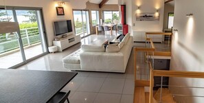 Ein modernes Wohnzimmer mit einem großen Flachbildfernseher, einem weißen Sofa, einem Essbereich mit Tisch und Stühlen und einer Treppe, die zu einer oberen Ebene führt.