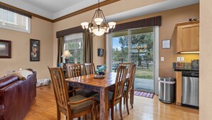 Un comedor con una mesa de madera, sillas y una araña. Hay una zona de cocina con lavavajillas y un cubo de basura.