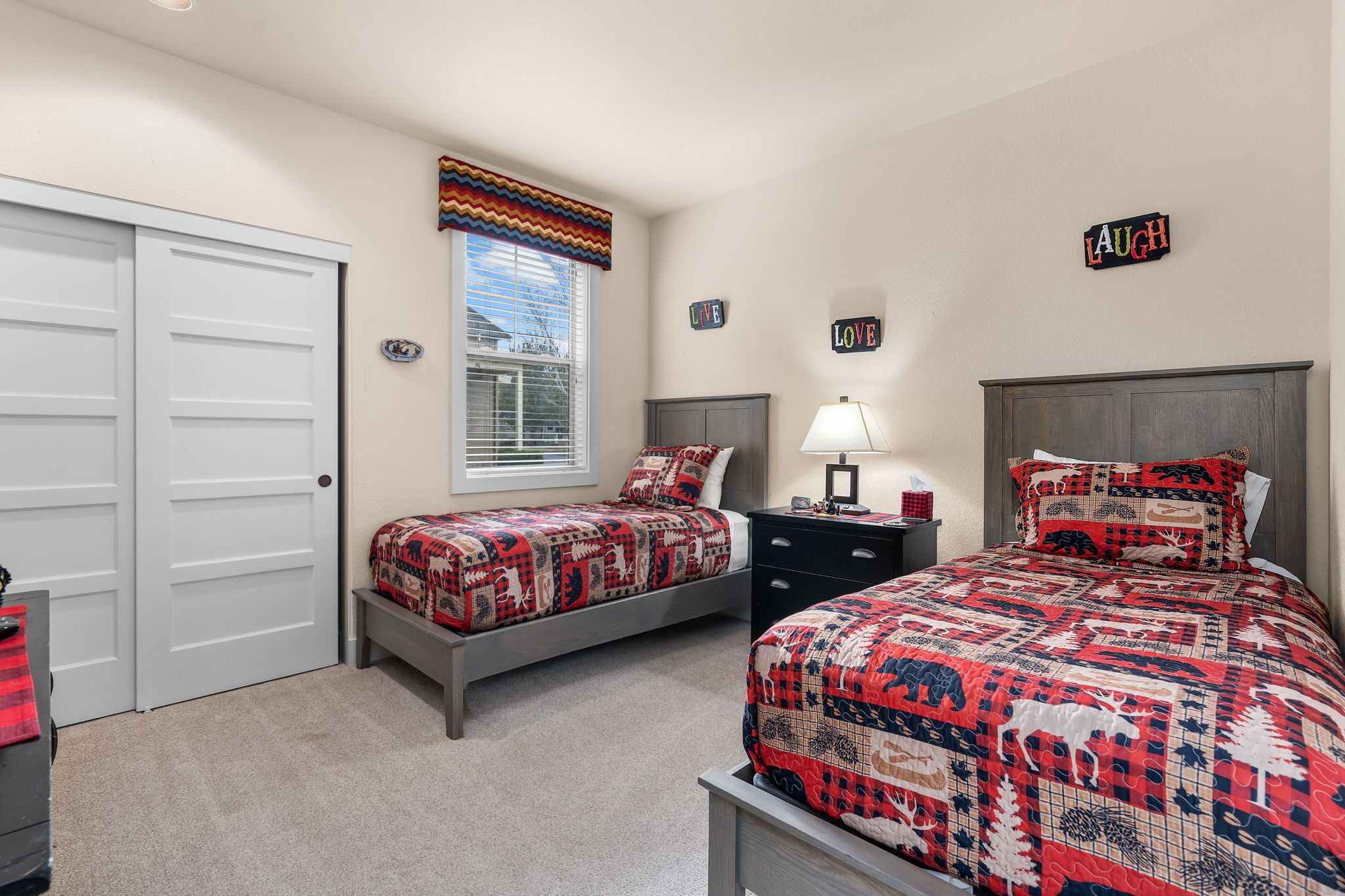 A bedroom with two beds, a window with blinds, a door, and a nightstand with a lamp.