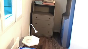 Ein kleines Zimmer mit einem Bett, einem Nachttisch mit Lampe, einer Kommode und einem Fenster mit Jalousien.