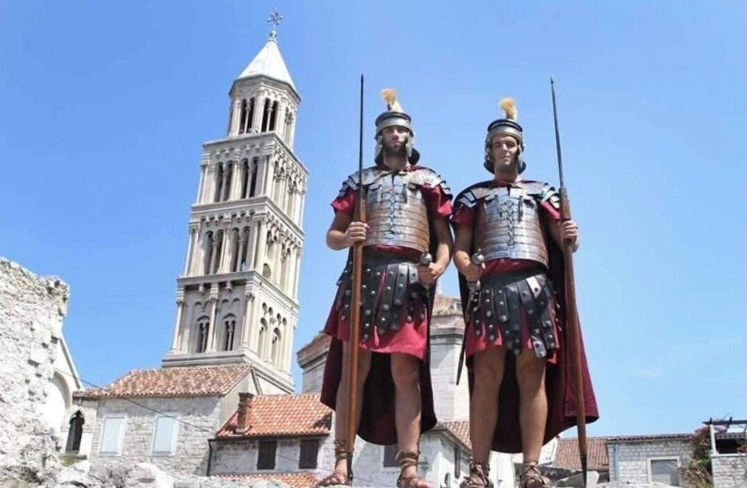Twee personen in Romeins harnas staan voor een historisch gebouw met een klokkentoren.