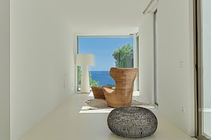 Ein modernes Interieur mit einem Korbstuhl, einem runden Hocker und Meerblick durch eine Glastür.