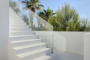 Eine weiße Treppe mit Glasgeländern, die zu einem Balkon mit Blick auf Palmen und weitere Grünflächen führt.